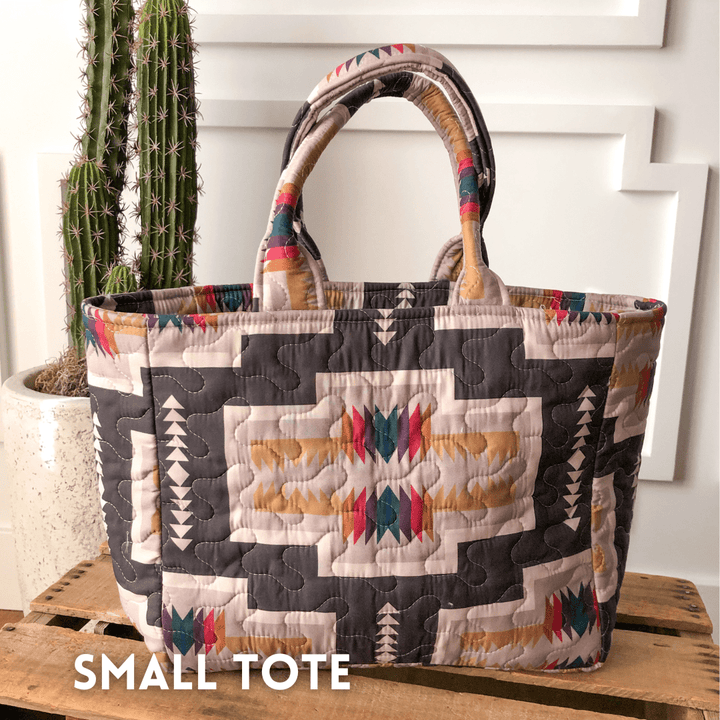 A quilted bag from The Muskogee Collection by Savannah Sevens western life{&}style, featuring colorful geometric patterns in beige, black, and red, rests on a wooden surface beside a potted cactus; SMALL TOTE appears in the lower left. By Ida Mae. 