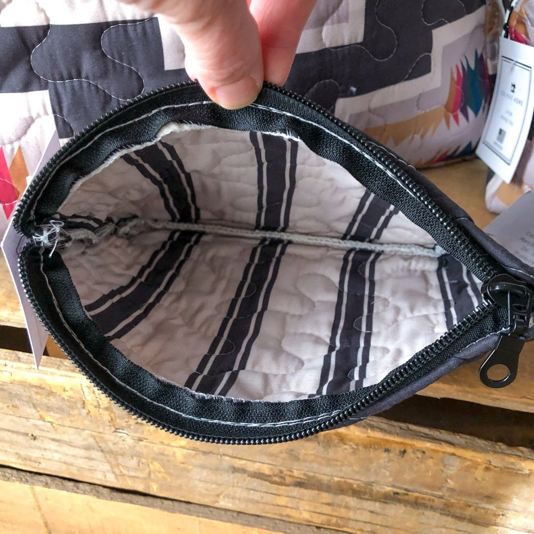 A hand opens The Muskogee {Collection} zippered pouch by Savannah Sevens western life{&}style, showing its striped quilted lining and empty interior. The bag sits on a wooden surface, with other patterned fabrics in the background. By Ida Mae. 
