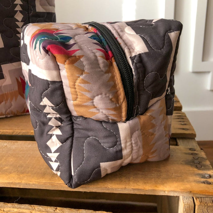 The Muskogee {Collection} quilted bag by Savannah Sevens western life{&}style features a black zipper and a colorful southwest design in brown, beige, red, and green with wavy stitching, displayed on a wooden surface. By Ida Mae. 