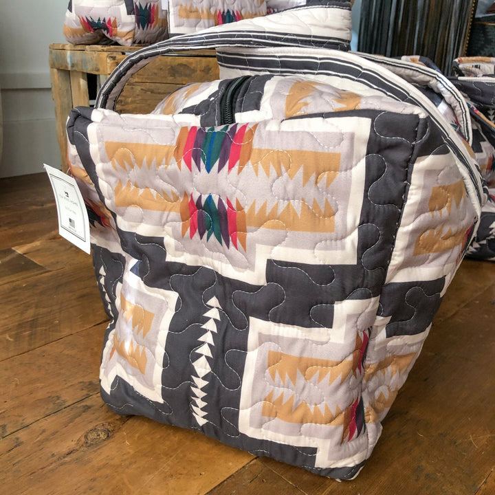 A bag from The Muskogee Collection by Savannah Sevens western life&style, featuring a southwest geometric pattern in beige, gray, and red with a black zipper and white tag, sits on a wooden floor among similar western bags. By Ida Mae. 