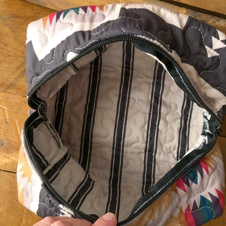 A hand holds open The Muskogee {Collection} bag by Savannah Sevens western life{&}style, featuring a colorful geometric exterior, black zipper, and a white quilted interior with black and gray vertical stripes. By Ida Mae. 