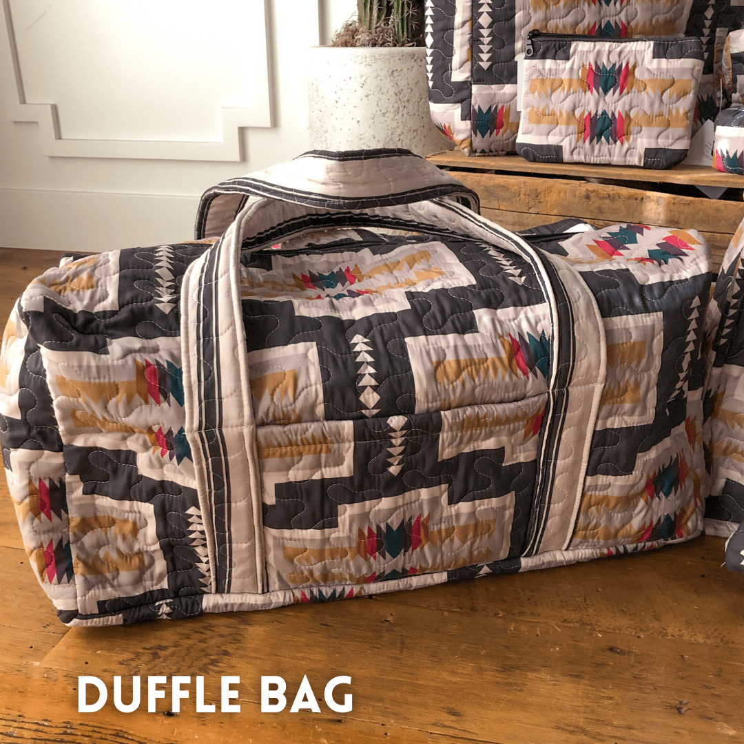 A colorful geometric bag from The Muskogee {Collection} by Savannah Sevens western life{&}style rests on a wooden floor, with similar bags in the background. "DUFFLE BAG" is written in white at the bottom left. By Ida Mae. 