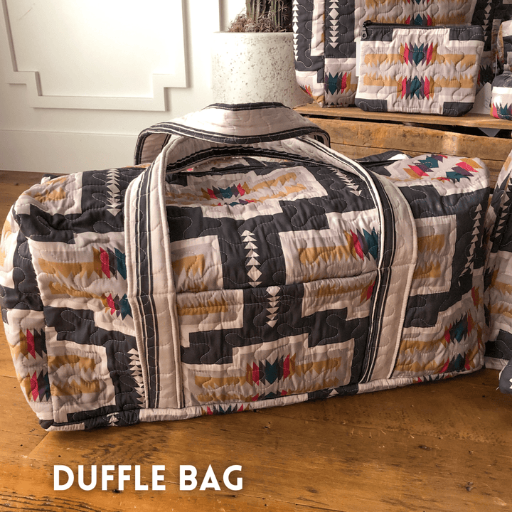 A colorful geometric bag from The Muskogee {Collection} by Savannah Sevens western life{&}style rests on a wooden floor, with similar bags in the background. "DUFFLE BAG" is written in white at the bottom left. By Ida Mae. 