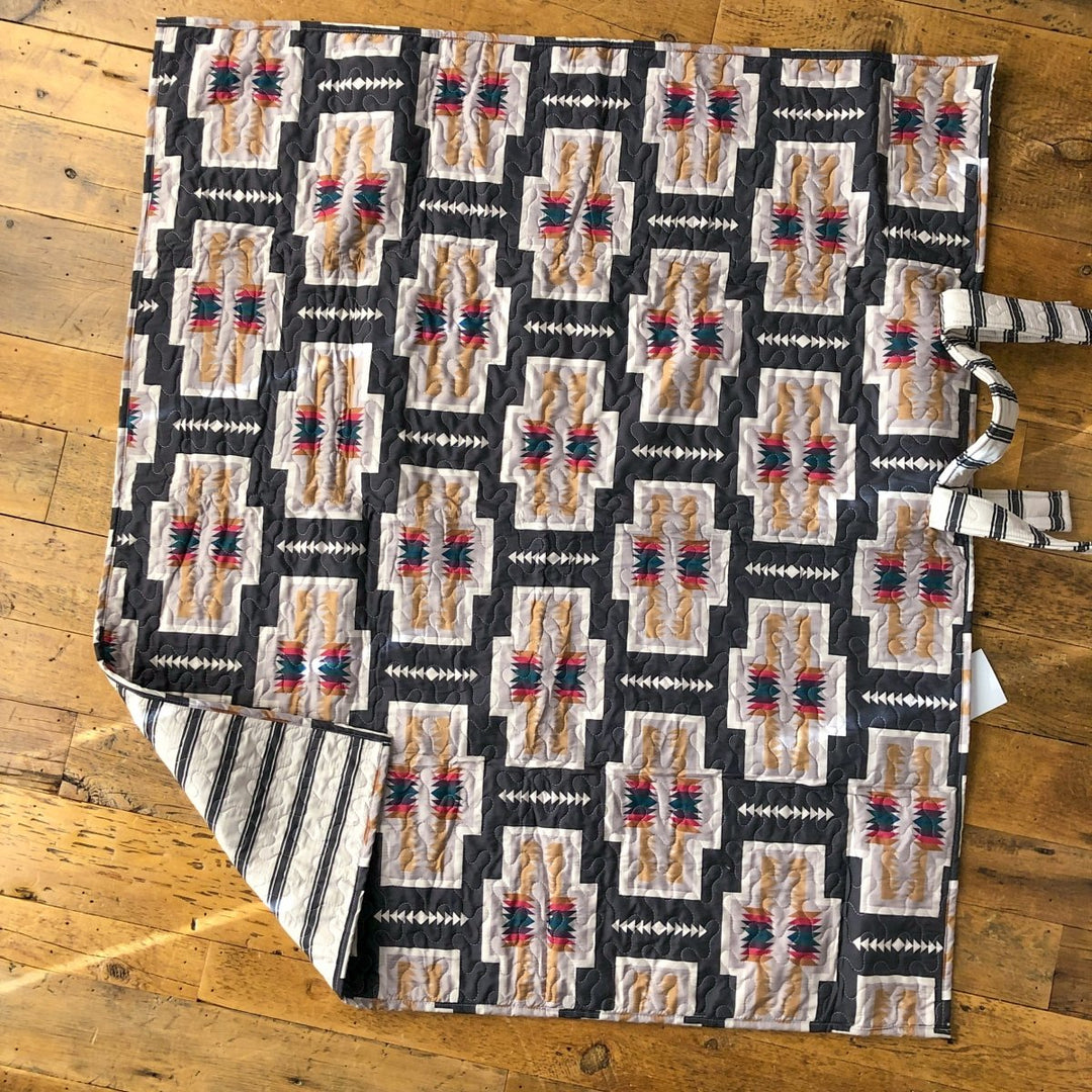 A quilt from The Muskogee {Collection} by Savannah Sevens western life{&}style, featuring geometric patterns in gray, beige, black, and red, lies on a wooden floor with striped backing and fabric ties visible on one side. By Ida Mae. 