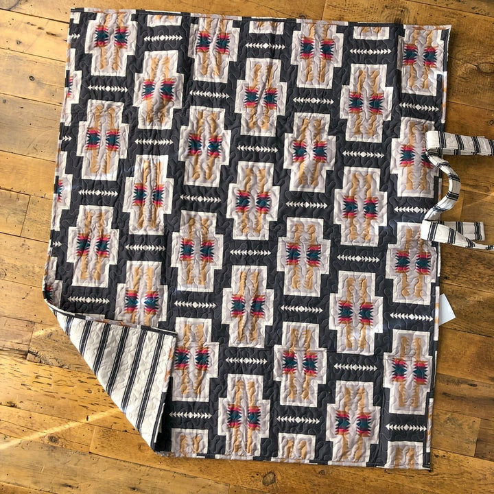 A quilt from The Muskogee {Collection} by Savannah Sevens western life{&}style, featuring geometric patterns in gray, beige, black, and red, lies on a wooden floor with striped backing and fabric ties visible on one side. By Ida Mae. 