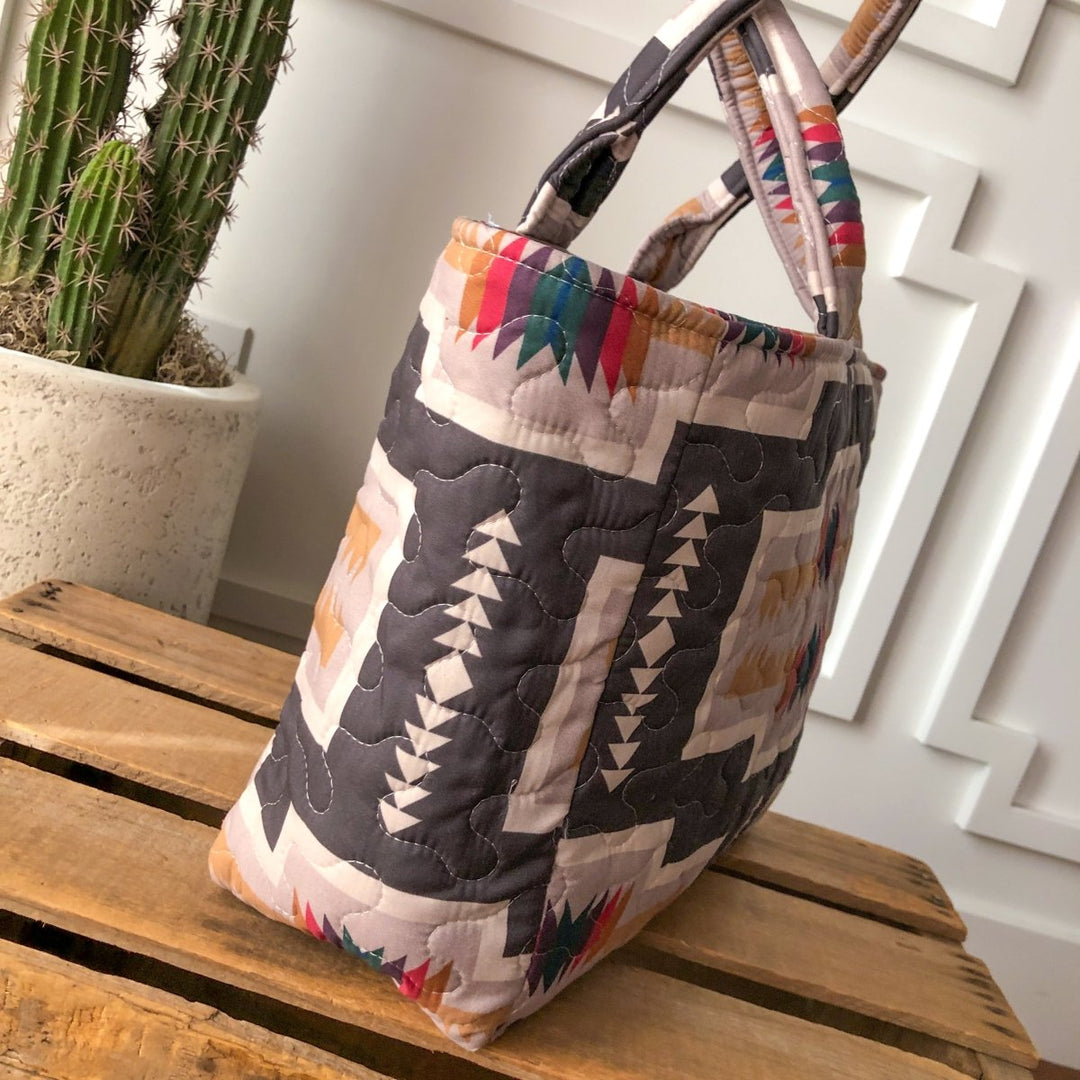 The Muskogee {Collection} bag by Savannah Sevens western life{&}style, with southwest geometric and tribal patterns in neutral and vibrant hues, is displayed on a wooden crate beside a potted cactus against a white textured wall. By Ida Mae. 