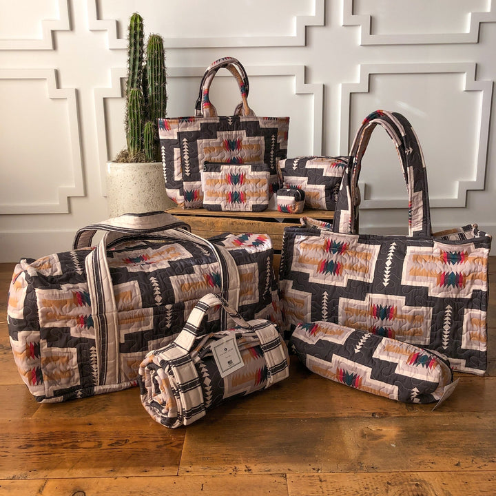 The Muskogee Collection by Savannah Sevens western life&style features geometric Southwestern-style quilted bags—Ida Mae totes, duffels, and small western bags—displayed on a wooden floor with a white paneled wall and potted cactus. By Ida Mae. 