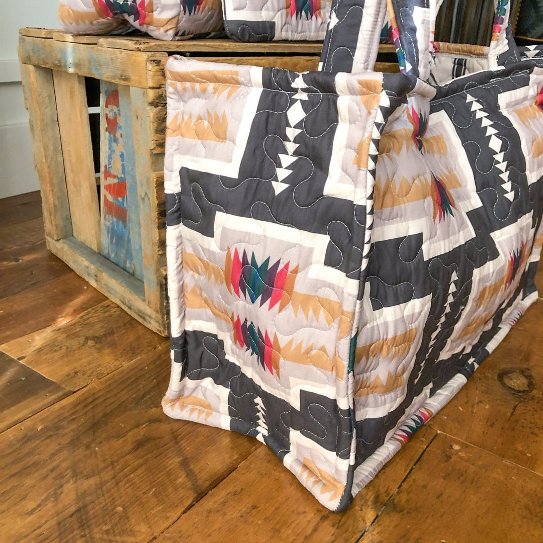 The Muskogee Collection quilted bag by Savannah Sevens western life&style, featuring southwest geometric patterns in black, beige, white, pink, and teal, rests on a wooden floor beside a rustic crate.