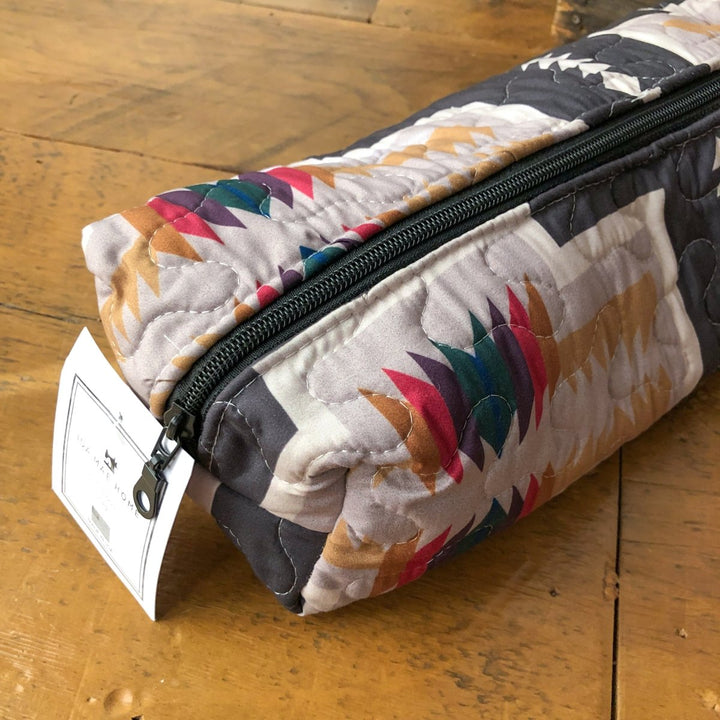 A close-up of The Muskogee {Collection} quilted bag by Savannah Sevens western life{&}style, featuring a black zipper, white tag, and geometric patterns in red, purple, green, orange, and beige on a wooden surface. By Ida Mae. 