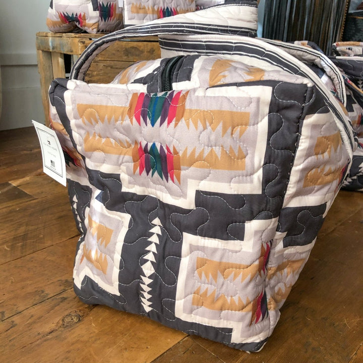 A bag from The Muskogee Collection by Savannah Sevens western life&style, featuring a southwest geometric pattern in beige, gray, and red with a black zipper and white tag, sits on a wooden floor among similar western bags. By Ida Mae. 
