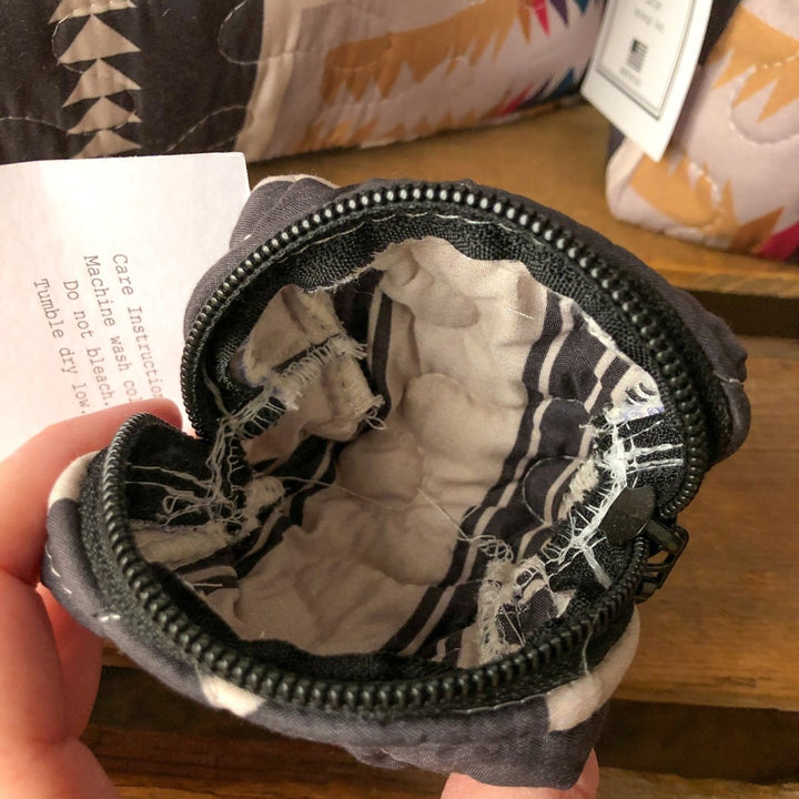 A close-up of The Muskogee {Collection} small, round quilted bag by Savannah Sevens western life{&}style shows its black zipper open, revealing white and black fabric interior with a care tag partially visible inside. By Ida Mae.