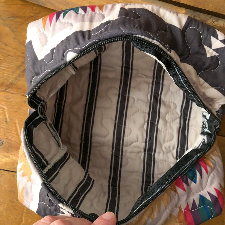 A hand holds open The Muskogee {Collection} bag by Savannah Sevens western life{&}style, featuring a colorful geometric exterior, black zipper, and a white quilted interior with black and gray vertical stripes. By Ida Mae. 