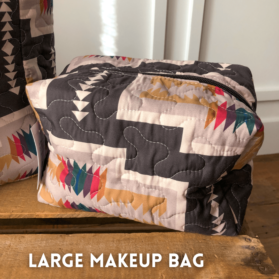 A large quilted bag from The Muskogee {Collection} by Savannah Sevens western life{&}style, featuring a colorful geometric pattern and black zipper, is shown on a wooden surface with "LARGE MAKEUP BAG" at the bottom. By Ida Mae. 
