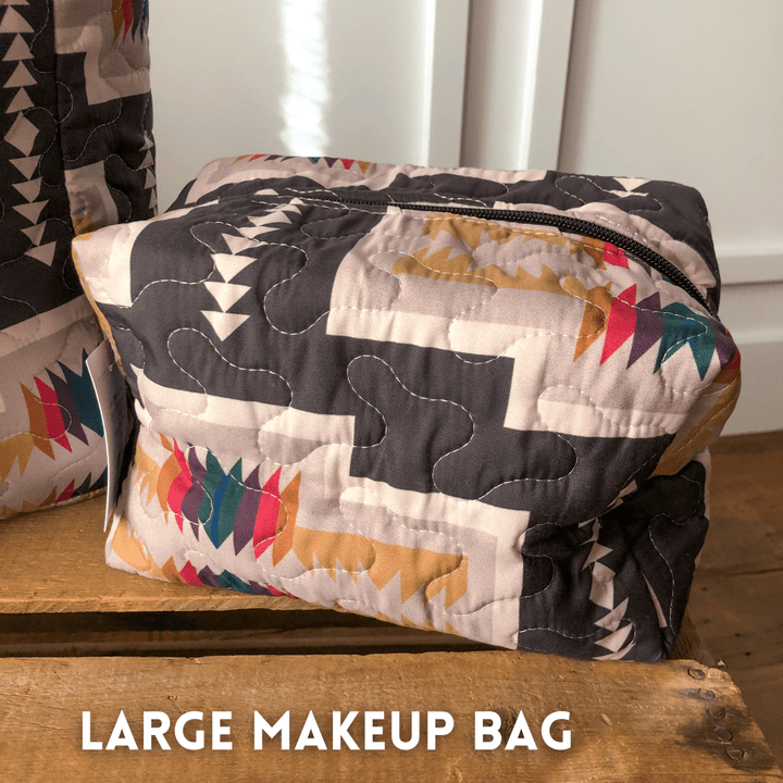 A large quilted bag from The Muskogee {Collection} by Savannah Sevens western life{&}style, featuring a colorful geometric pattern and black zipper, is shown on a wooden surface with "LARGE MAKEUP BAG" at the bottom. By Ida Mae. 