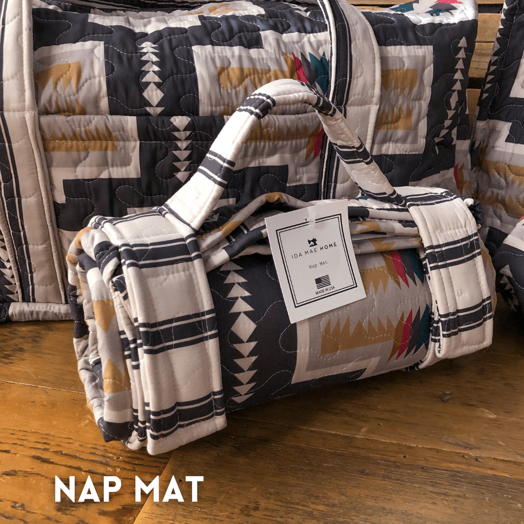 A rolled-up nap mat from The Muskogee Collection by Savannah Sevens western life&style, featuring a southwest-inspired geometric pattern, sits on a wooden floor with handles, an IDA MAT HOME tag, and "NAP MAT" in the bottom left corner.