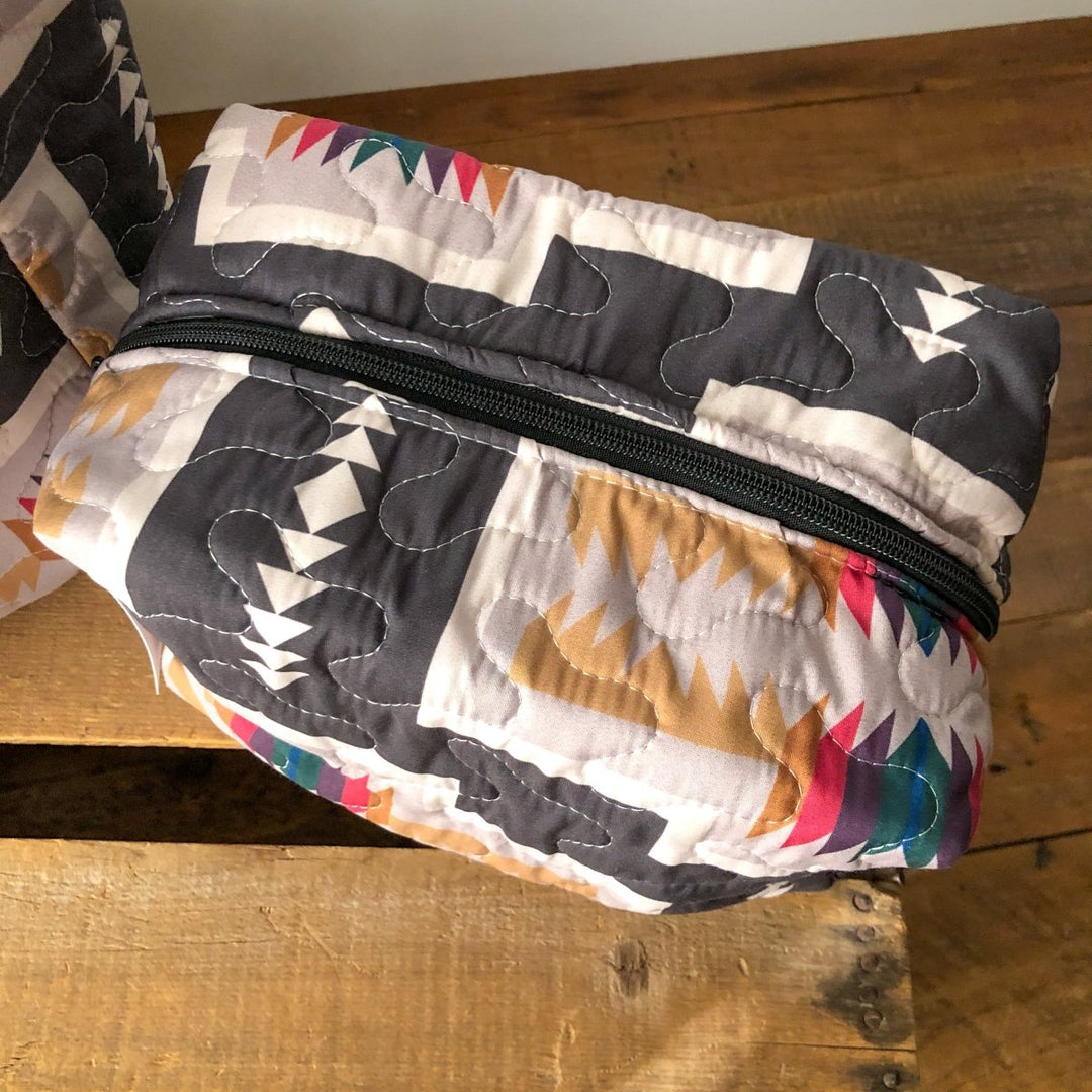 The Muskogee Collection quilted bag by Savannah Sevens western life&style, featuring southwest geometric patterns in black, white, brown, and colorful accents, is displayed on a wooden surface. By Ida Mae.