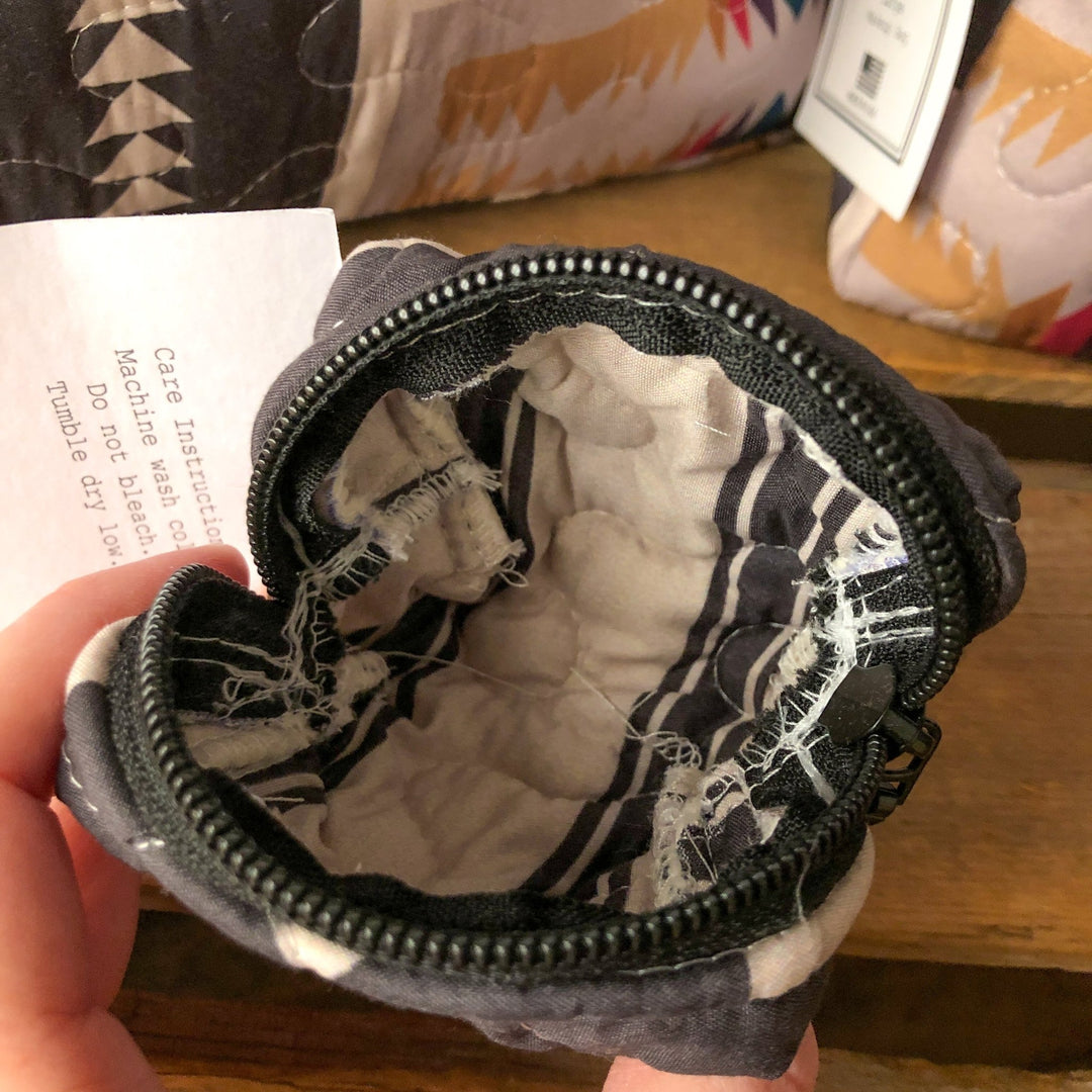A close-up of The Muskogee {Collection} small, round quilted bag by Savannah Sevens western life{&}style shows its black zipper open, revealing white and black fabric interior with a care tag partially visible inside. By Ida Mae.