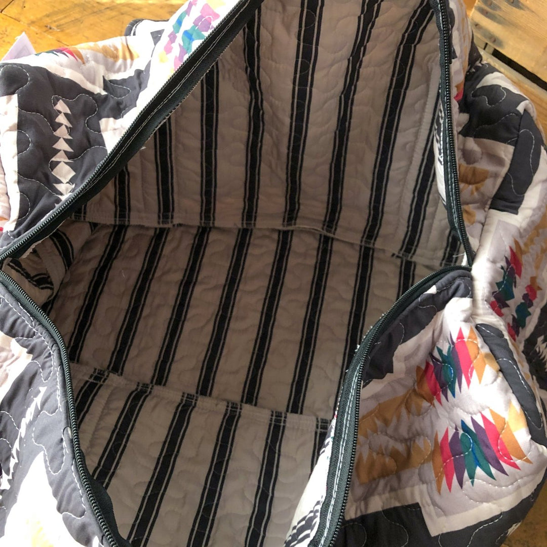 The Muskogee {Collection} quilted bag by Savannah Sevens western life{&}style displays colorful geometric and gray patterns, with a striped lining visible through the zippered top, shown on a wooden surface. By Ida Mae. 