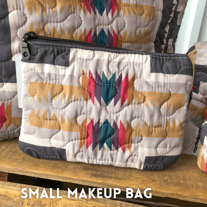 A small zippered bag from The Muskogee Collection by Savannah Sevens western life&style, featuring a colorful southwest geometric quilted pattern, stands on wood. "SMALL MAKEUP BAG" appears in white at the image's bottom. By Hooey. 