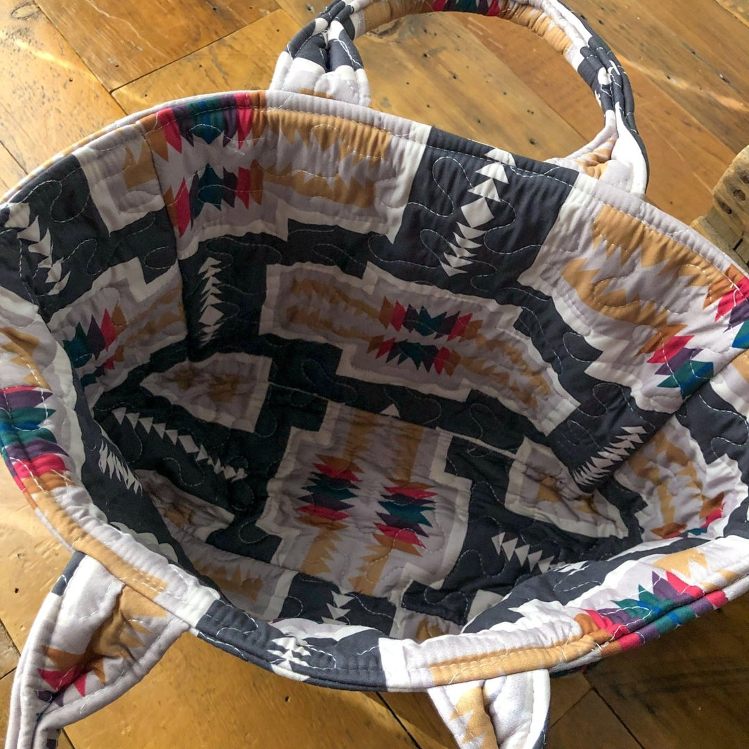 The Muskogee Collection bag by Savannah Sevens western life&style features a colorful southwest geometric pattern in black, tan, red, and teal, and sits open on a wooden floor displaying its unique interior design. By Ida Mae.