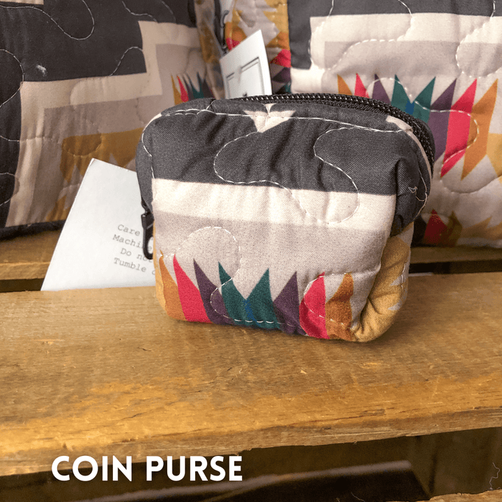 A small, quilted coin purse from The Muskogee Collection by Savannah Sevens western life{&}style features a colorful geometric pattern. It sits on a wooden surface with a top zipper and a partially visible tag behind it. By Ida Mae.