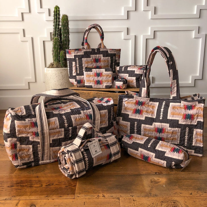 The Muskogee Collection by Savannah Sevens western life&style features geometric Southwestern-style quilted bags—Ida Mae totes, duffels, and small western bags—displayed on a wooden floor with a white paneled wall and potted cactus. By Ida Mae. 