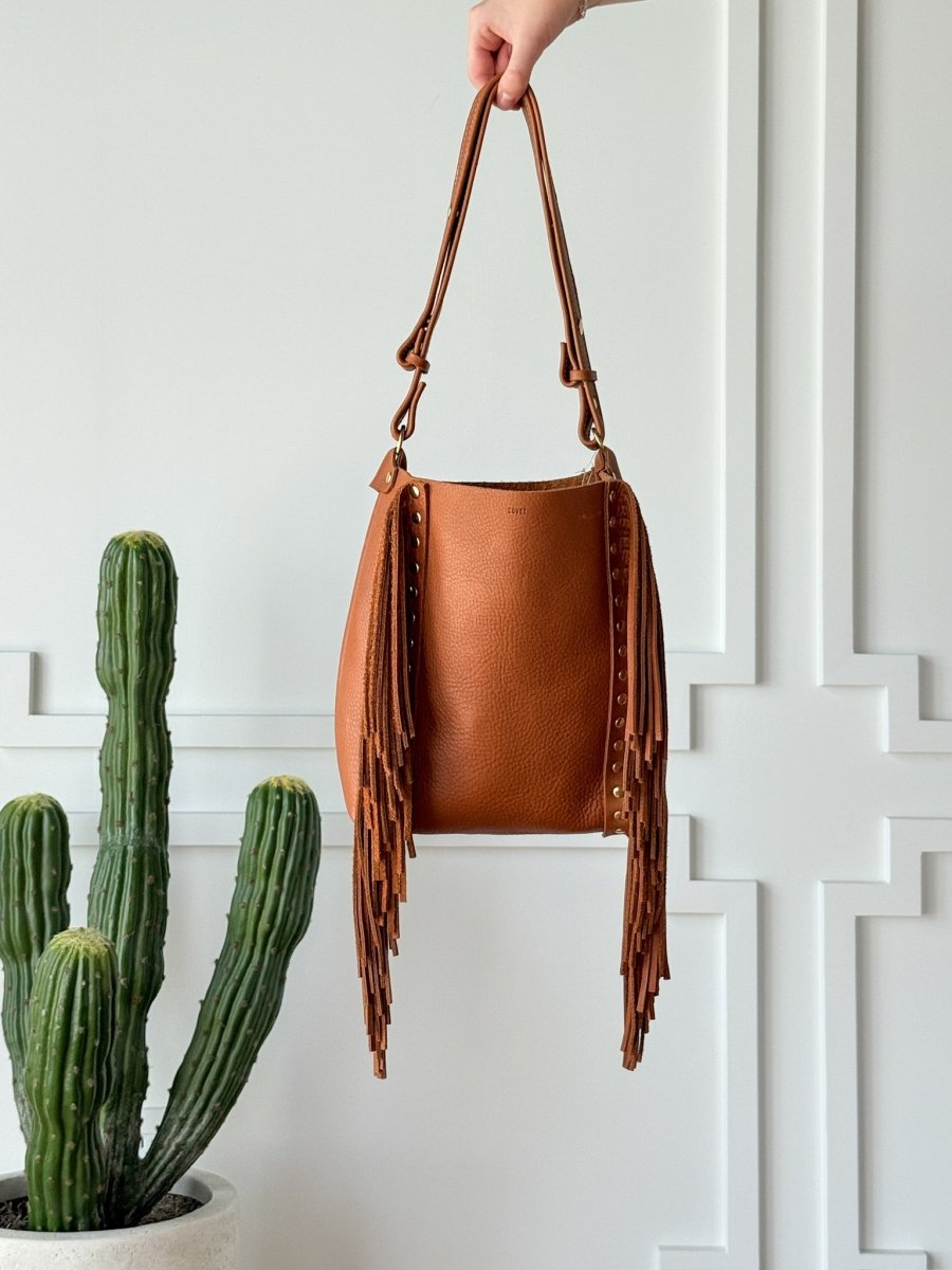 A hand holds up The Navasota full-grain leather fringe handbag by Savannah Sevens western life{&}style, displayed next to a tall green cactus in a white pot against a white paneled wall. By Covet. 