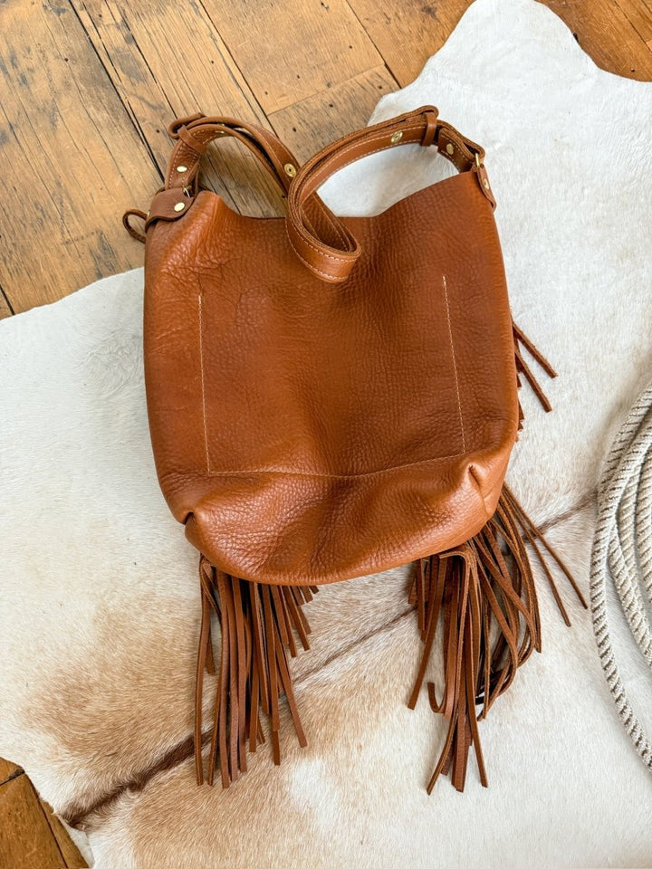 The Navasota by Savannah Sevens western life{&}style is a pecan full-grain leather tote with gold hardware and long fringe accents, styled on a light cowhide rug over wood, highlighting its textured surface and fine craftsmanship. By Covet. 