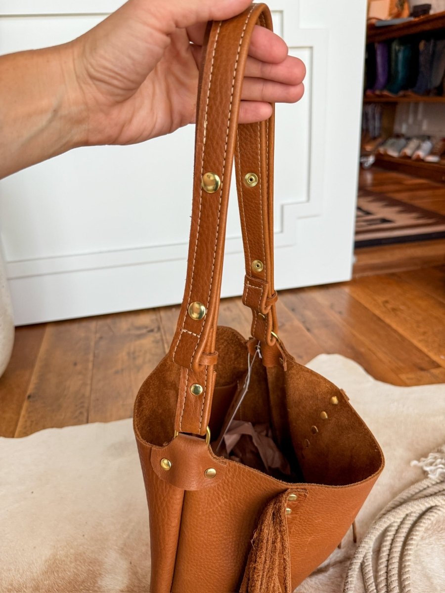 A hand holds The Navasota tote by Savannah Sevens western life{&}style, featuring pecan full-grain leather with gold rivets and a tassel, shown against a wooden floor and white cabinet background. By Covet. 