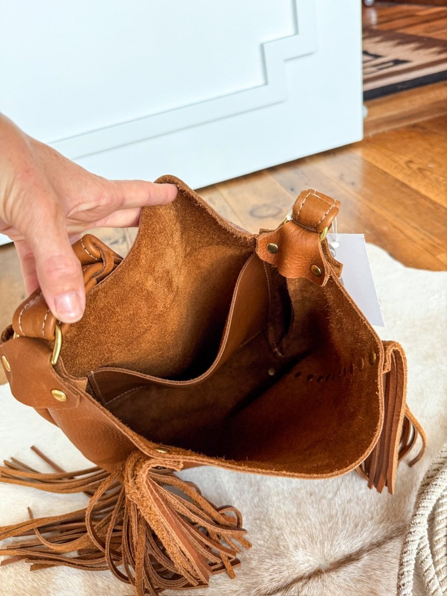 A hand opens The Navasota tote by Savannah Sevens western life{&}style, showing its suede interior and small inner pocket. The full-grain pecan leather bag sits on a cream surface with wooden flooring in the background. By Covet. 