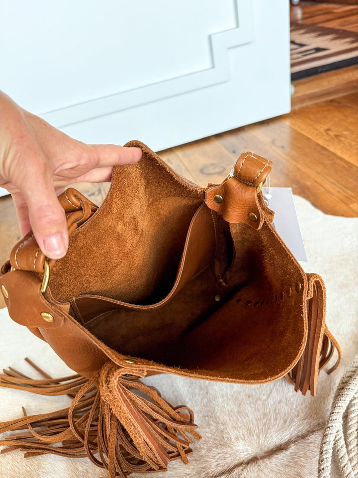 A hand opens The Navasota tote by Savannah Sevens western life{&}style, showing its suede interior and small inner pocket. The full-grain pecan leather bag sits on a cream surface with wooden flooring in the background. By Covet. 