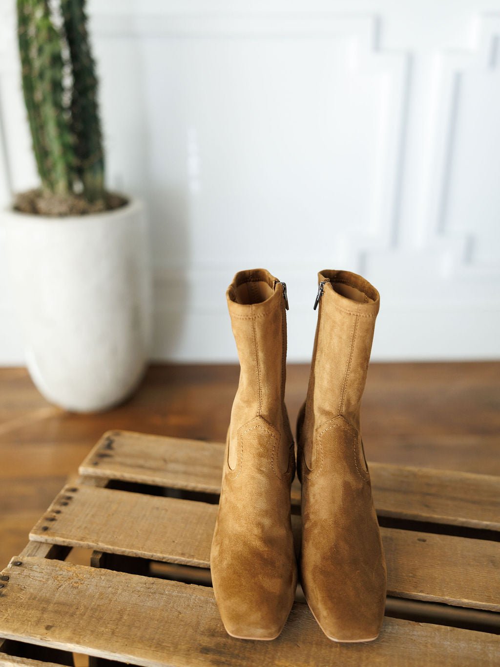 The Palmetto booties by Savannah Sevens western life{&}style, in tan faux suede with square toes, are displayed side by side on a wooden pallet. A tall potted cactus stands in the background against a white wall.