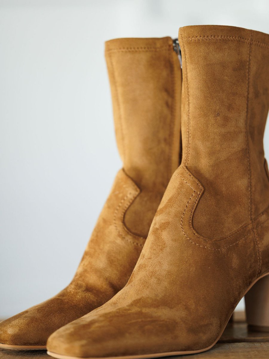 A close-up of The Palmetto tan faux suede booties by Savannah Sevens western life{&}style, featuring a rounded square toe, side zipper, and block heel, set on a light surface with a soft neutral background.
