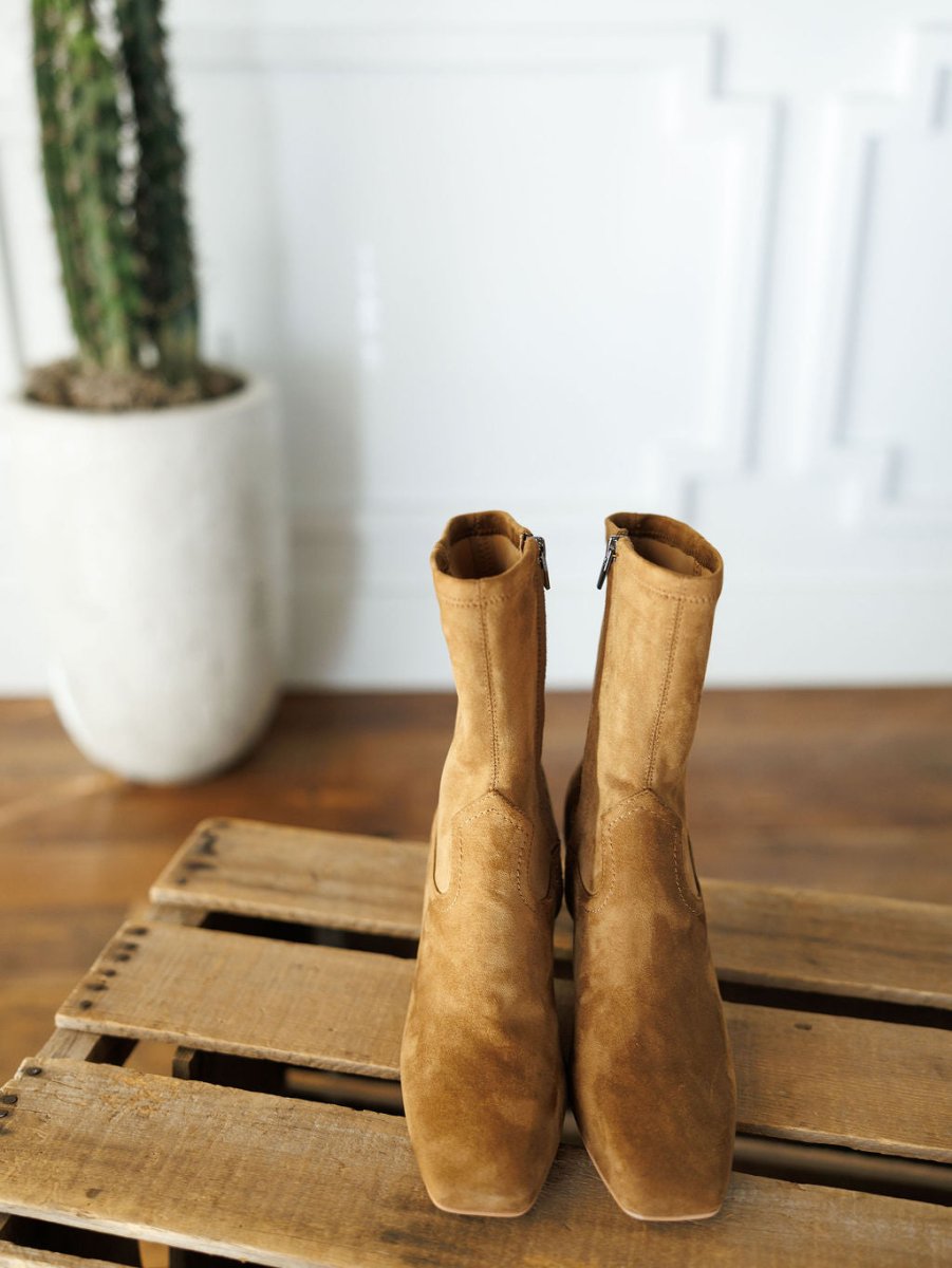 The Palmetto booties by Savannah Sevens western life{&}style, in tan faux suede with square toes, are displayed side by side on a wooden pallet. A tall potted cactus stands in the background against a white wall.