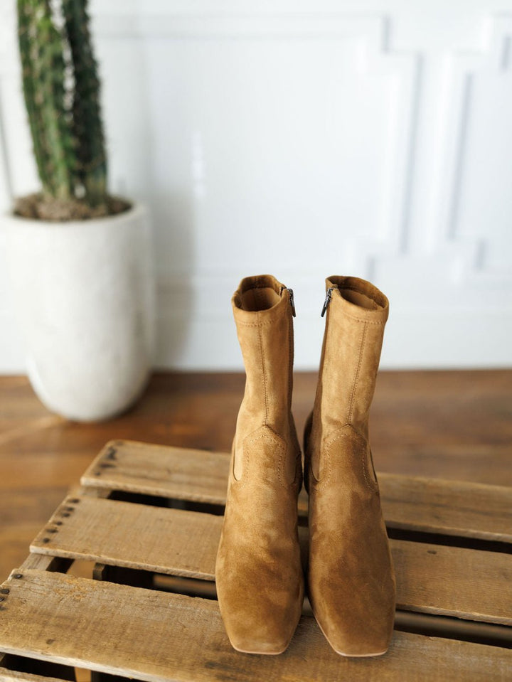 The Palmetto booties by Savannah Sevens western life{&}style, in tan faux suede with square toes, are displayed side by side on a wooden pallet. A tall potted cactus stands in the background against a white wall.