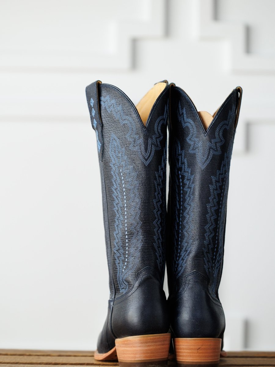 A pair of tall midnight navy leather "The Pasadena" boots by Savannah Sevens western life{&}style, featuring decorative stitching, stands side by side on a wooden surface against a white geometric-patterned background. Boots made by Blackstar.