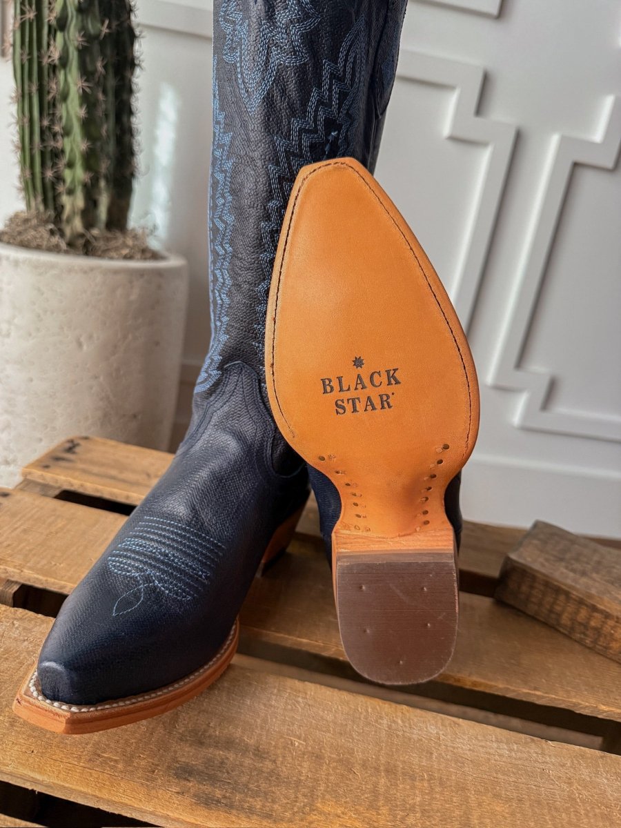 A close-up of a person wearing The Pasadena midnight navy leather western snip toe boot by Savannah Sevens western life{&}style, with the sole visible. They're standing on wood, and a tall cactus in a white pot is in the background. Boots by Blackstar. 