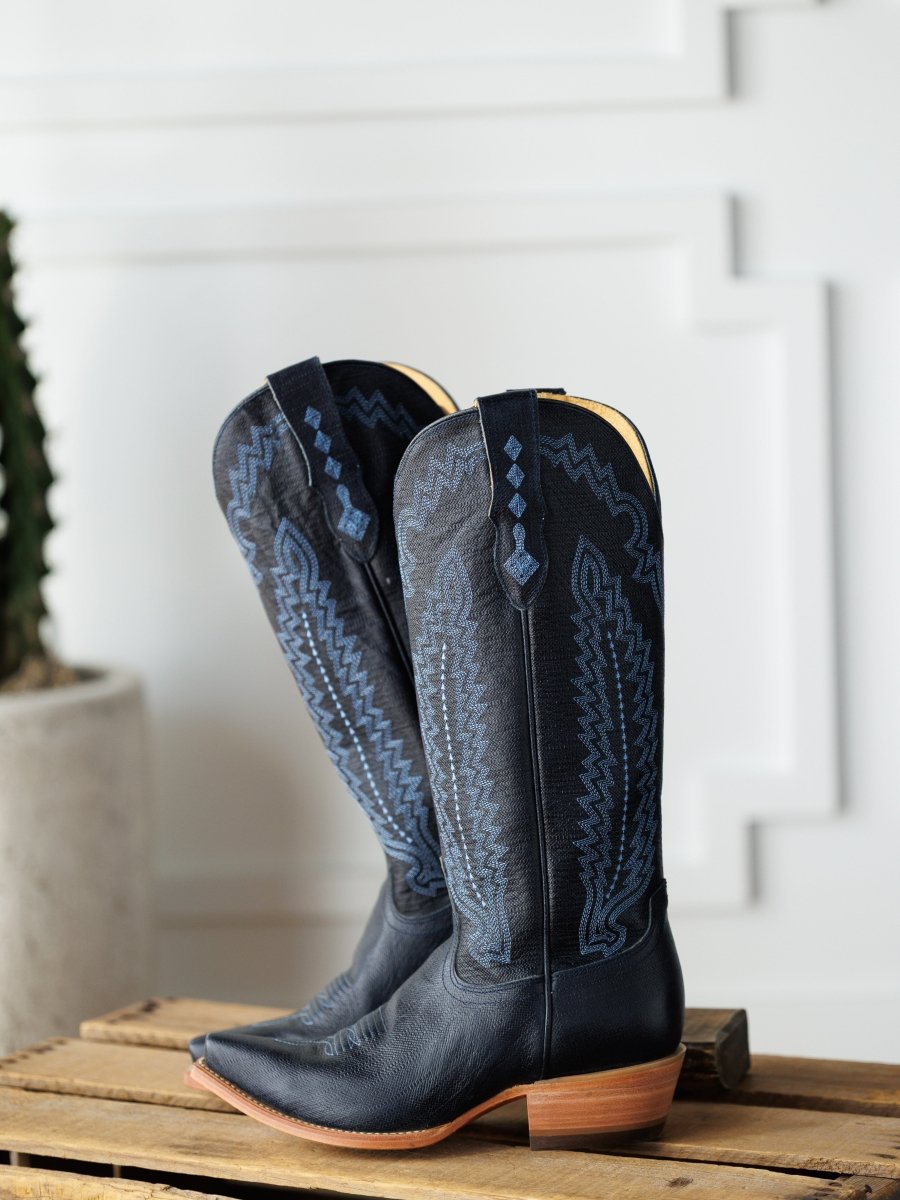 The Pasadena boots by Savannah Sevens western life{&}style, featuring black leather and blue embroidery, rest on a wooden surface with a white textured wall and a cactus in the background. Boots made by Blackstar.
