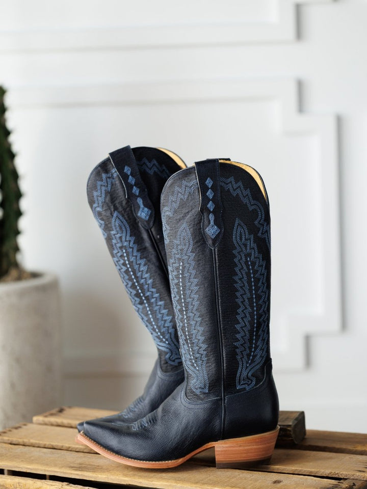 The Pasadena boots by Savannah Sevens western life{&}style, featuring black leather and blue embroidery, rest on a wooden surface with a white textured wall and a cactus in the background. Boots made by Blackstar.