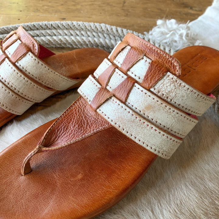 Close-up of Savannah Sevens western life{&}style’s The Pensacola distressed leather sandal with woven straps, resting on a light furry surface. Another sandal and a coiled rope appear in the background.