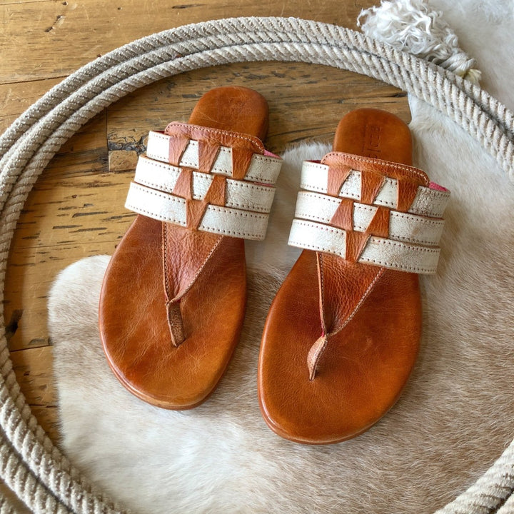 The Pensacola sandals by Savannah Sevens western life{&}style, featuring distressed leather and cream woven straps, are displayed on a cowhide rug with coiled rope nearby atop a wooden floor.