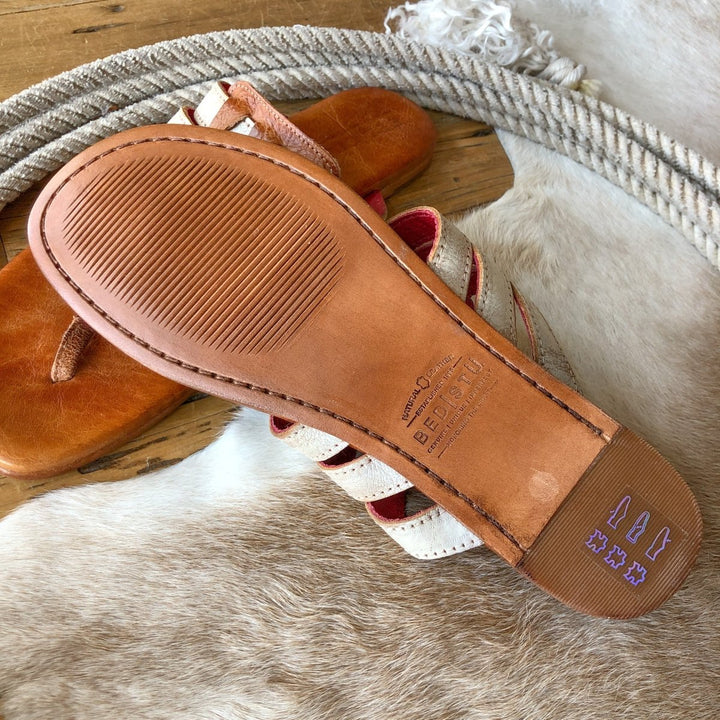 A close-up of the sole of The Pensacola sandal by Savannah Sevens western life{&}style on a light animal hide rug, with another woven strappy sandal and a coiled rope in the background. Branding and textured tread are visible on the sole. Sandals made by BedStu. 