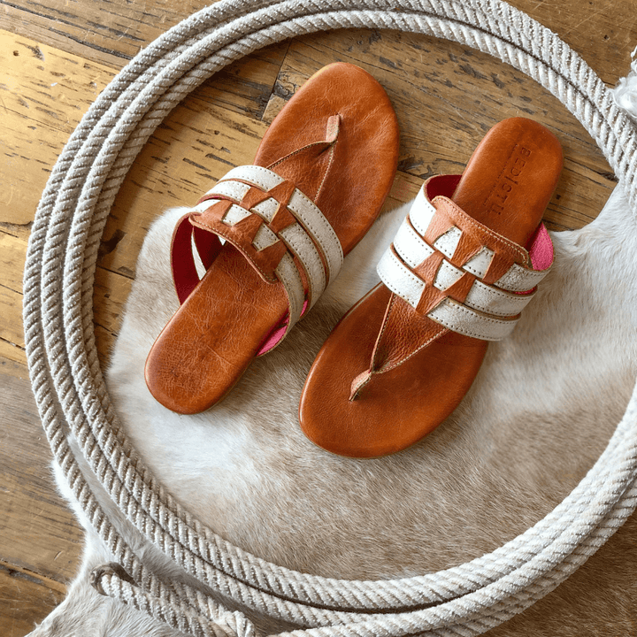 A pair of The Pensacola tan and white woven strappy sandals by Savannah Sevens western life{&}style rests on a cowhide rug with a coiled rope nearby, atop a wooden floor.