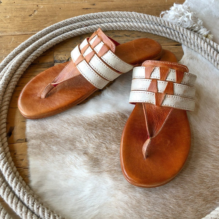The Pensacola sandals by Savannah Sevens western life{&}style feature distressed leather and wide cream and tan woven straps, shown on a cowhide rug inside a coiled rope atop a wooden floor.