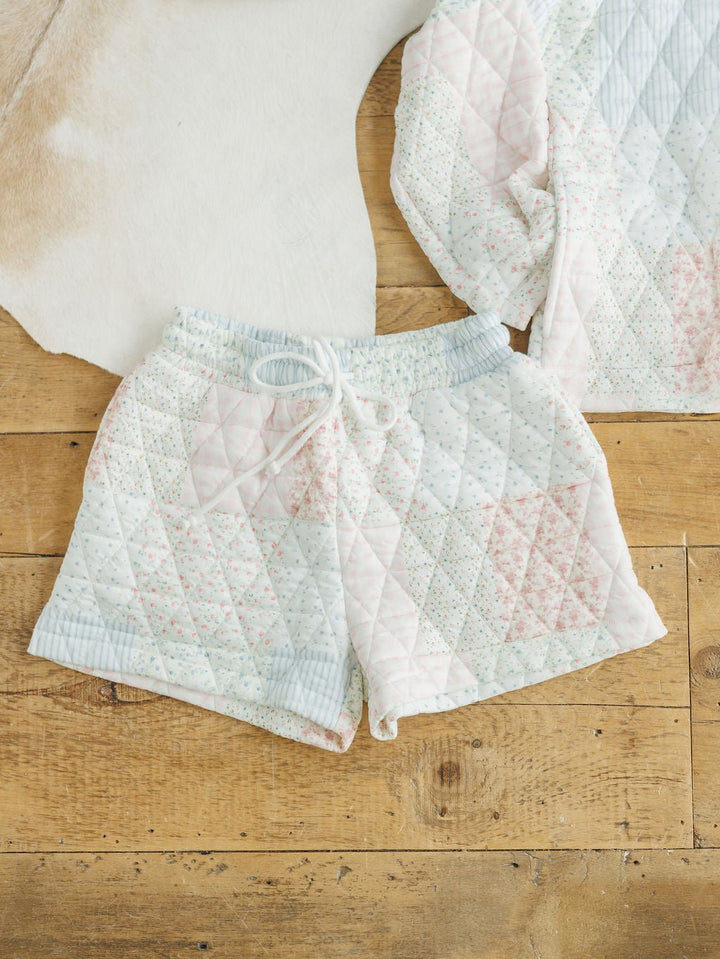 The Petaluma {Set} by Savannah Sevens western life{&}style features high-waisted, relaxed-fit quilted pajama shorts in a pastel patchwork pattern with a white drawstring, shown with the matching top on a wooden floor by a white rug.