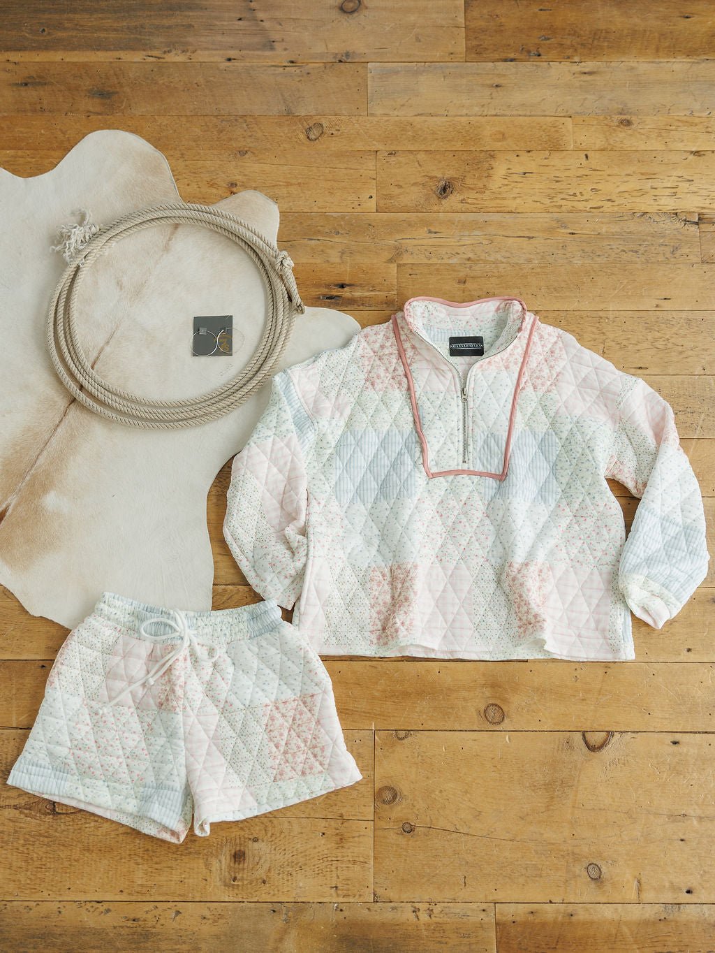 The Petaluma {Set} by Savannah Sevens western life{&}style—a pastel quilted patchwork pullover with a zip collar and relaxed fit, plus high-waisted shorts—rests on a wooden floor beside a cream cowhide rug and coiled rope.