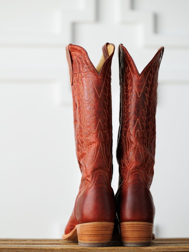 The Pueblo boots by Savannah Sevens western life{&}style feature tall, brown distressed leather with decorative stitching, shown upright on a wooden surface against a white background.