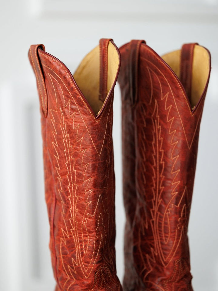 A close-up of two "The Pueblo" boots by Savannah Sevens western life{&}style, made from distressed leather with detailed stitching, standing side by side against a softly blurred background.