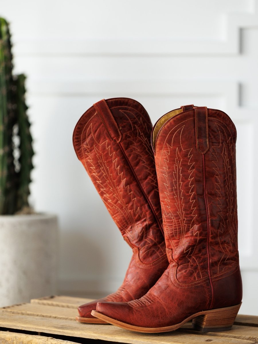 The Pueblo boots by Savannah Sevens western life{&}style are tall, distressed leather western boots with intricate stitching, displayed on wood with a blurred potted cactus in the background. Boots made by Blackstar.