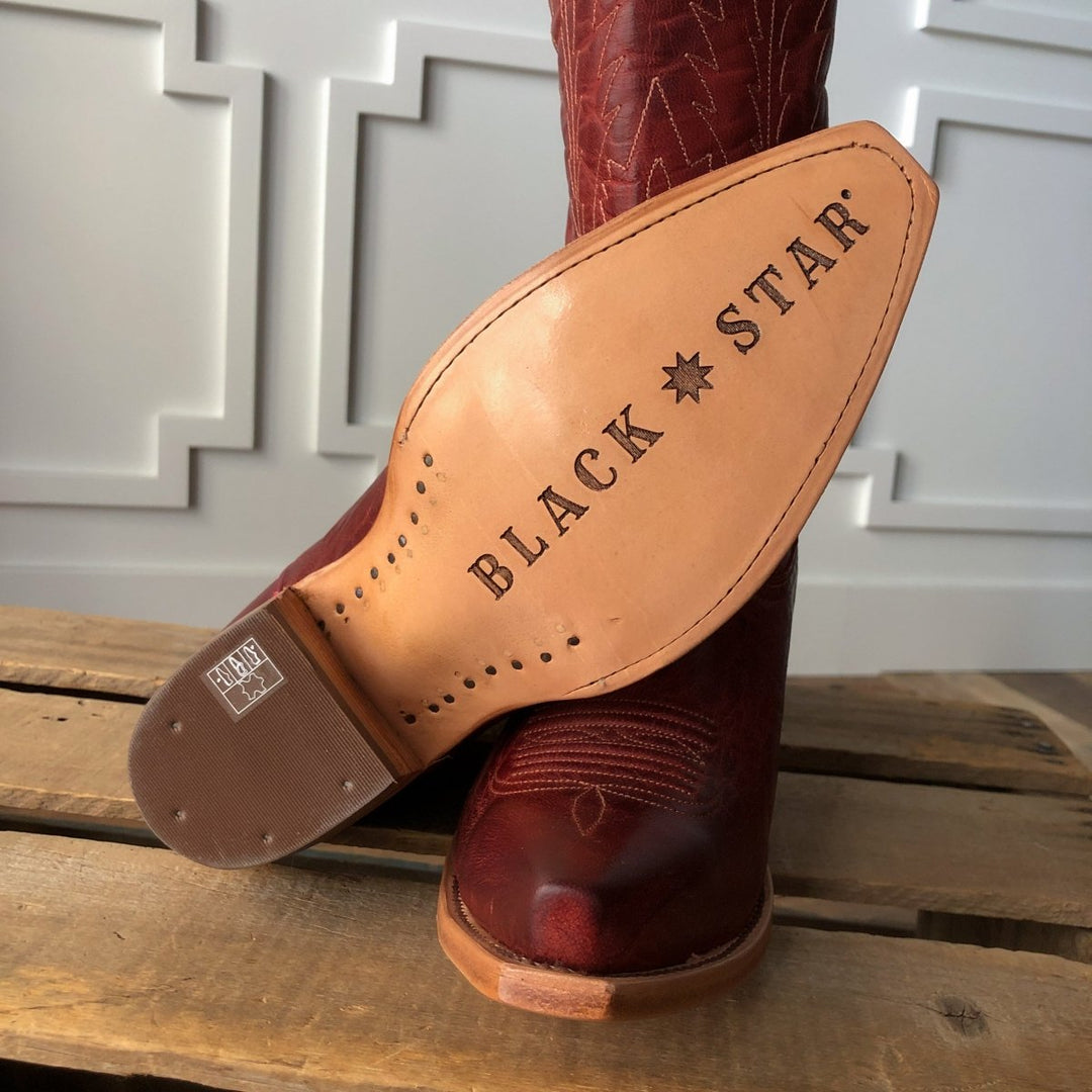 A close-up of The Pueblo boot sole by Savannah Sevens western life{&}style shows its stamped logo on distressed leather. This burgundy boot features stitching and sits on a wooden surface in front of a white paneled wall. Boots made by Black Star. 
